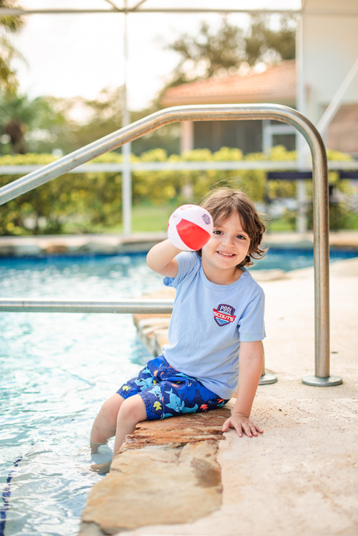 Little boy sitting along edge of pool holding ball and smiling