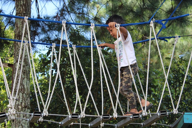 Little boy walking across a rope bridge on a playground with trees in background