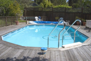 Swimming pool surrounded by wooden deck in backyard with fence and trees behind it