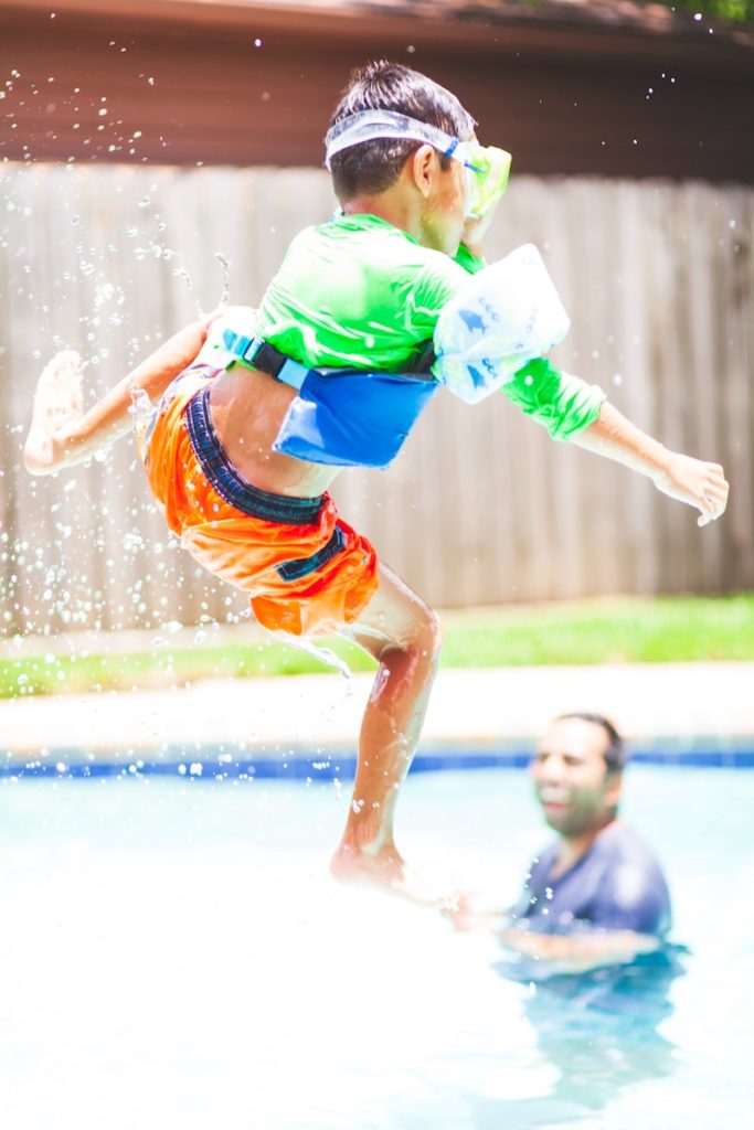 Image of little boy getting thrown in the air in a pool with floaties and goggles, and dad in the background